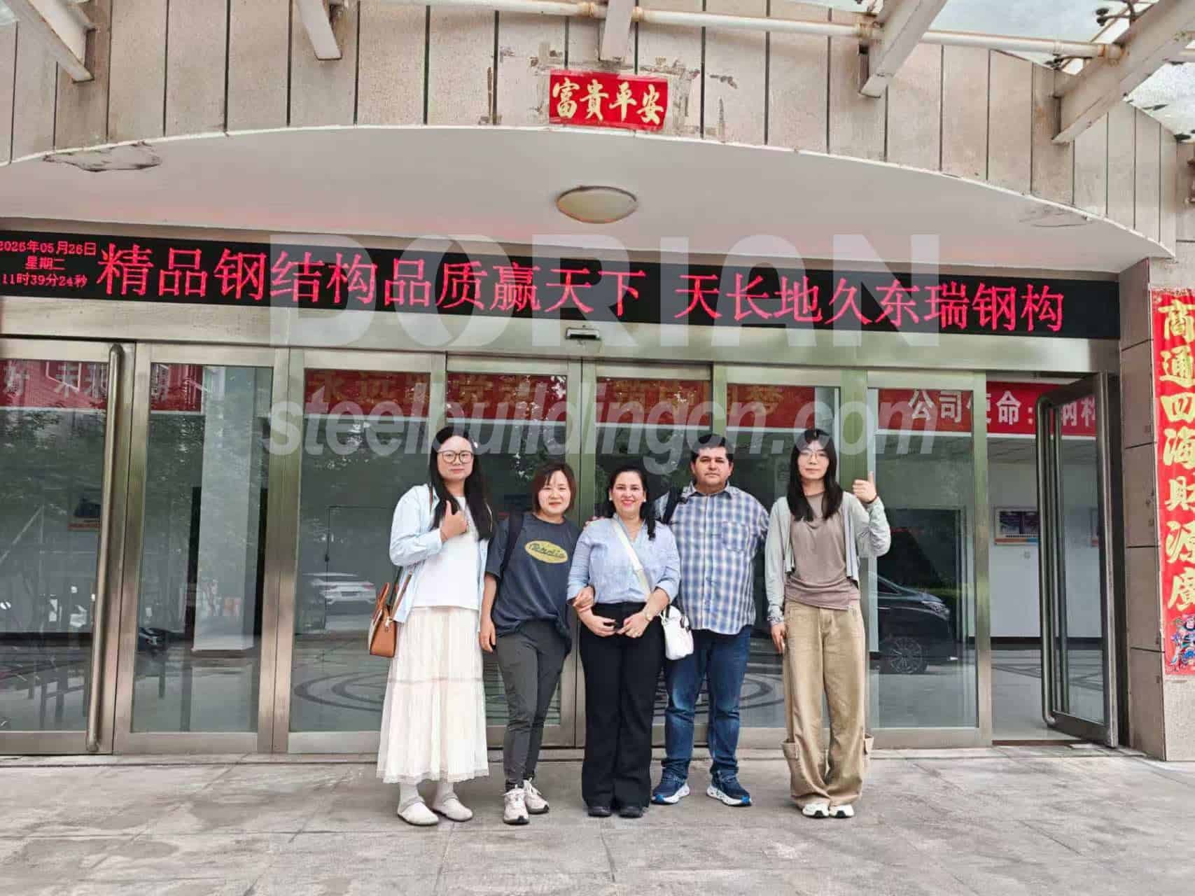 Group of people standing in front of a steel building entrance with glass doors and signage. Modern steel building with glass entrance and signage
